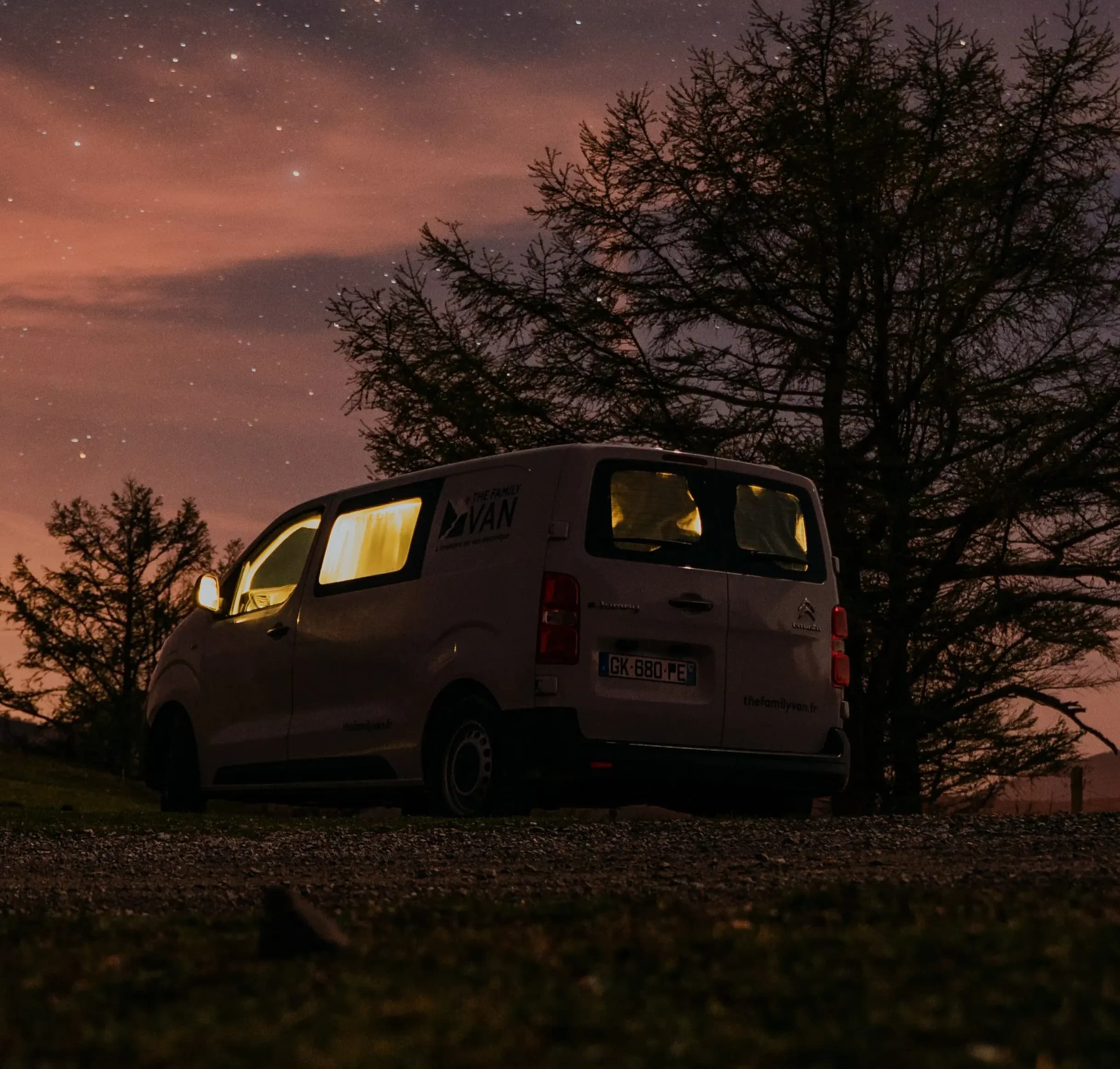 Campervan éclairé de l'intérieur sous un ciel étoilé, silhouettes d'arbres tout autour