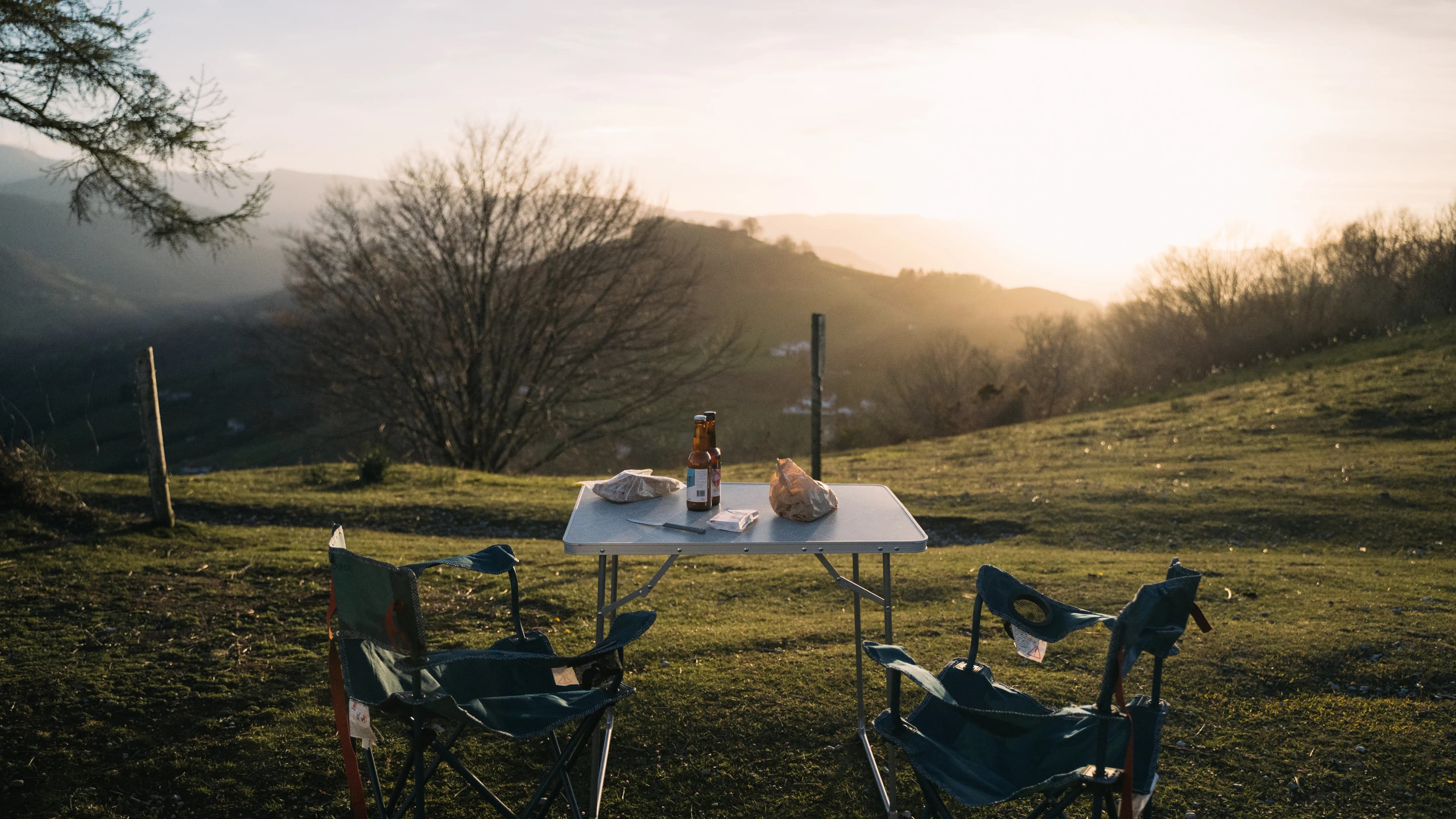 Table de camping dressée avec une bière et deux chaises pliantes face aux collines au crépuscule